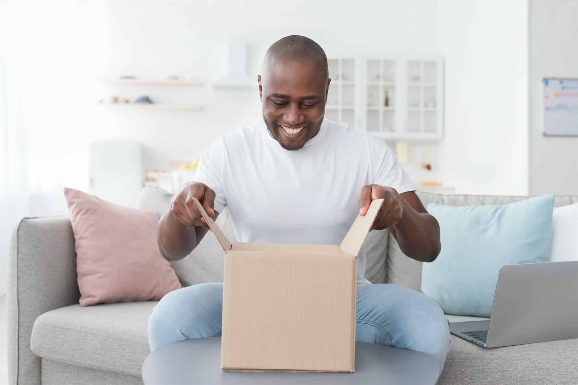 Happy black man unpacking delivery box, sitting on couch at home interior. Satisfied customer