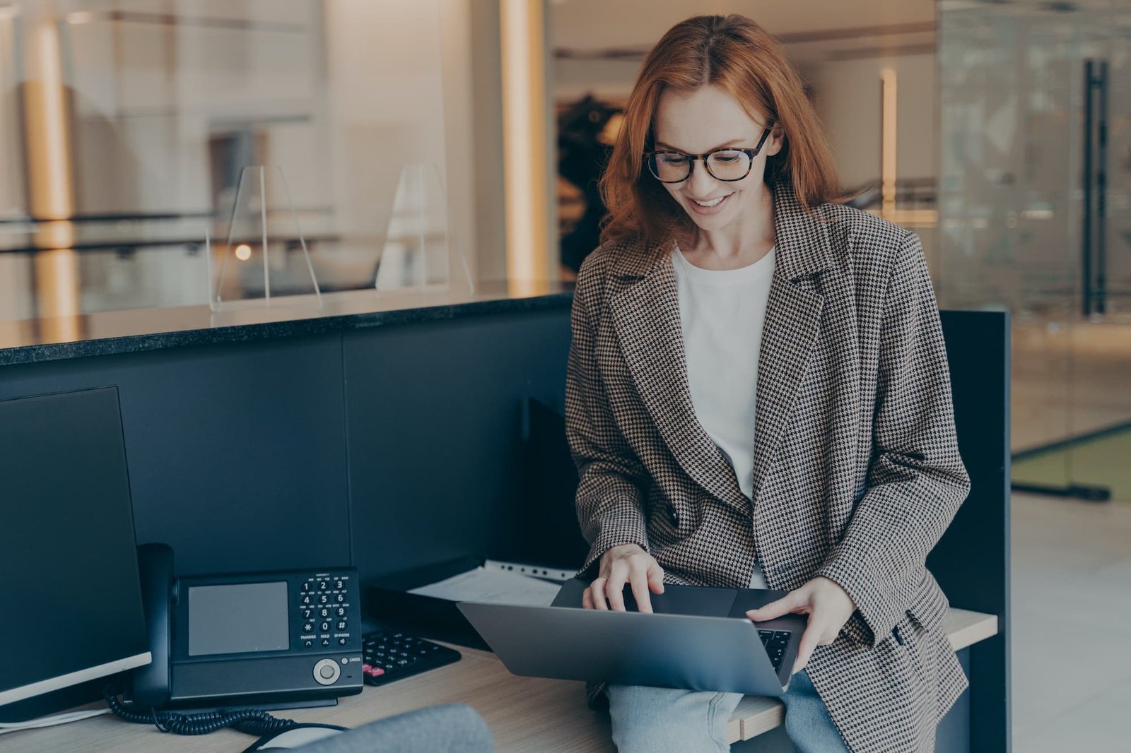 Beautiful redhead businesswoman sitting on top of desk and using laptop, working alone in office